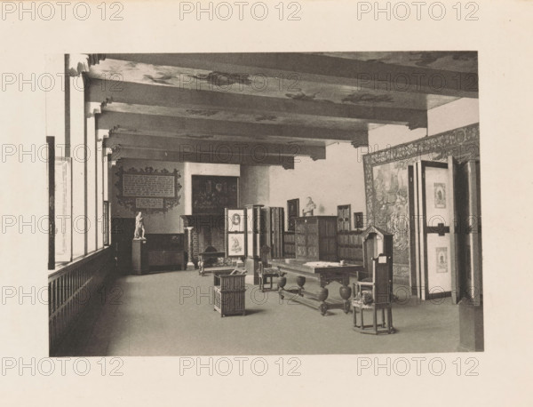 Refectory of the former St. Agatha Monastery in Delft, page, photomechanical print, title: 'Refectory of the Convent of St. Agatha, Delft.', photographer: Charles L. Mitchell, Prinsenhof (Delft), cliché maker: AW Elson & Company, Boston (Massachusetts), c. 1884 - in or before 1894, Delft, North America, Massachusetts, paper, photoaquatint, height 87 mm x width 124 mm, interior, representation of a building, refectory, monastery, A long interior room with exposed ceiling beams and patterned ceiling panels, a row of tall windows along the left side, wooden wainscoting beneath the windows, and a decorative framed panel on the far wall. Various pieces of wooden furniture are arranged in the space including tables, chairs, and cabinets, a folding screen with framed images, and a pedestal topped by a small statue. A large tapestry or mural covers the right wall near an open doorway, and smaller framed artworks are displayed on the walls and atop furniture. The floor is open and mostly clear ...
