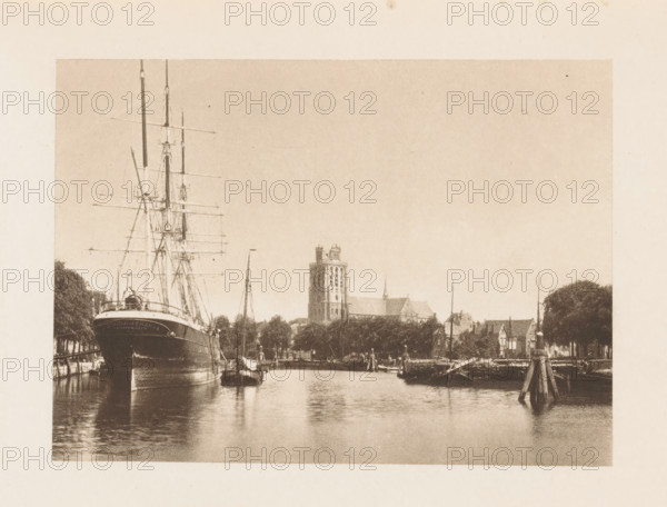 View of the Meuse at Dordrecht, with the Grote or Onze-Lieve-Vrouwekerk in the background, page, photomechanical print, title: 'Dordrecht-Canal with Cathedral in the Distance.', photographer: Charles L. Mitchell, Dordrecht, clichémaker: AW Elson & Company, Boston (Massachusetts), c. 1884 - in or before 1894, North America, Massachusetts, paper, photoaquatint, height 88 mm x width 123 mm, river, church (exterior), ships (in general), Grote or Onze-Lieve-Vrouwekerk, A sepia-toned waterfront scene showing a large ship with tall masts moored at the left edge, its bow facing forward and rigging lines visible, smaller boats alongside and reflections in the calm water; a riverside town stretches across the middle distance with trees along the bank, a prominent multi-story tower and church spire rising above lower buildings, wooden posts and piers extending into the water at right, and an expanse of pale sky above.