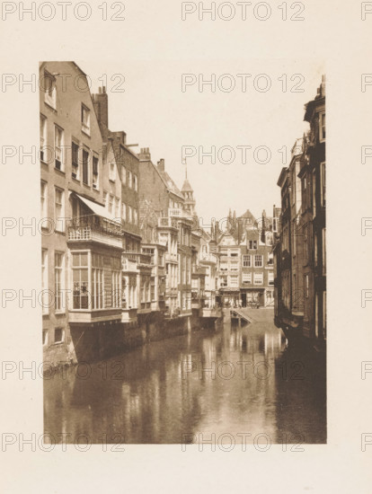 View of Het Steiger in Rotterdam, page, photomechanical print, title: 'The Steiger, Rotterdam', photographer: Charles L. Mitchell, Rotterdam, cliché maker: AW Elson & Company, Boston (Massachusetts), c. 1884 - in or before 1894, North America, Massachusetts, paper, photoaquatint, height 118 mm x width 86 mm, canals, waters (in city), facade (of house or building), Steiger, A narrow canal flanked by tall, closely packed buildings with multiple windows and decorative facades runs into the distance, the water reflecting the buildings and appearing smooth and dark, left-side structures showing bay windows, balconies, and awnings projecting over the canal while right-side buildings form a darker vertical edge, and in the background a cluster of ornate rooflines and a small tower rise above the street-level architecture.