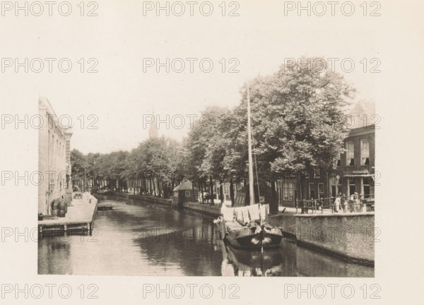 View of a canal in Delft, page, photomechanical print, title: 'Near the Arsenal, Delft.', photographer: Charles L. Mitchell, Delft, cliché maker: AW Elson & Company, Boston (Massachusetts), c. 1884 - in or before 1894, North America, Massachusetts, paper, photoaquatint, height 87 mm x width 117 mm, canals, waters (in city), A sepia-toned scene shows a narrow canal lined with buildings and trees, with a sailboat moored in the foreground reflecting in the water; a row of trees extends along the canal bank, and small boats and houseboats are visible along the water; on the right a quay with a railing borders buildings with windows and awnings, and a few people stand near the railing; on the left a dock area with barrels and steps leads to the canal, and a distant church spire rises above the treeline.