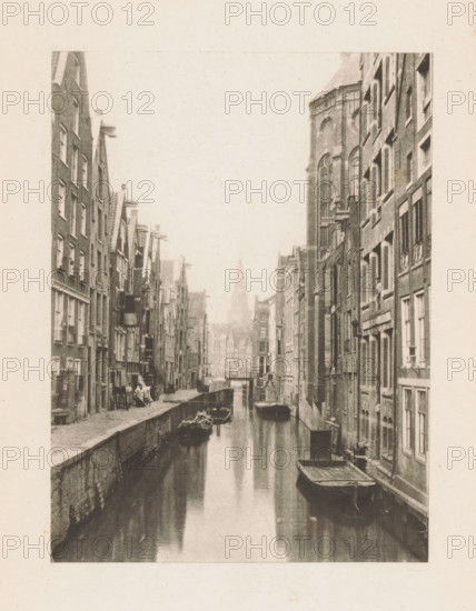 Canal in Amsterdam, page, photomechanical print, title: 'Old Amsterdam.', photographer: Charles L. Mitchell, Amsterdam, cliché maker: AW Elson & Company, Boston (Massachusetts), c. 1884 - in or before 1894, North America, Massachusetts, paper, photoaquatint, height 120 mm x width 88 mm, Print on frontispiece., canals, waters (in city), A narrow canal runs between tall brick buildings on both sides, with moored wooden boats floating on the water and a small bridge visible farther down the channel; the buildings show multiple windows and doors, some with shutters and decorative architectural details, and a larger building with tall arched windows rises prominently on the right; the canal edges have stone walkways and occasional small items and a few indistinct figures near the waterside; the scene is rendered in a sepia-toned, aged photographic style.