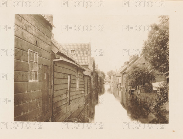 Houses along a ditch in Zaandam, page, photomechanical print, title: 'Afternoon in Zaandam.', photographer: Charles L. Mitchell, Zaandam, cliché maker: AW Elson & Company, Boston (Massachusetts), c. 1884 - in or before 1894, Zaanstreek, North America, Massachusetts, paper, photoaquatint, height 88 mm x width 125 mm, housing, canals, waters (in village), A narrow waterway runs down the center of the image, flanked by wooden buildings with slatted siding on the left and a mix of pitched roof houses and dense foliage on the right, their reflections visible in the water; the left structures show a small window and a door, and the rooftops display overlapping tiles and eaves, while the right side features trees and shrubs near the banks and a covered object near the edge.