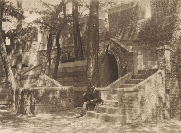 Inside of the Castle of Leiden, page, photomechanical print, title: 'Courtyard of the Burg, Leiden.', photographer: Charles L. Mitchell, Leiden, cliché maker: AW Elson & Company, Boston (Massachusetts), c. 1884 - in or before 1894, North America, Massachusetts, paper, photoaquatint, height 86 mm x width 116 mm, castle, walls, fortified city, Burcht van Leiden, A sepia-toned scene of a brick building with arched entryways and a flight of stone steps leading up to one of the openings, flanked by low brick walls and rounded stone structures; tall trees with textured trunks and leafy branches stand in front of the building, casting dappled shadows across the ground and walls; a man in dark clothing and a hat sits on the steps near the base of a tree, with one leg extended and one hand resting on his knee; the foreground shows a ground surface with scattered light patches and the image is bordered by a pale margin.