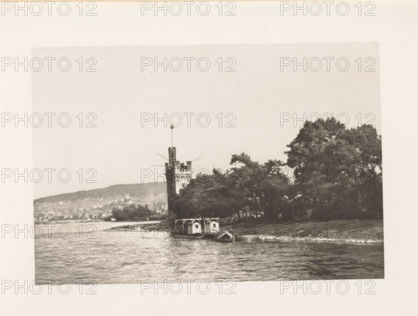 View of the Mouse Tower near Bingen am Rhein, page, photomechanical print, title: 'The Mouse Tower, Bingen.', Rhine, c. 1889 - in or before 1899, paper, photoaquatint, height 126 mm x width 87 mm, river, tower, clock tower, monumental adornment, Muizentoren, A riverside scene in a monochrome photograph shows a body of water in the foreground with rippling surface, a small boathouse structure moored at the shore with a narrow boat alongside, a grassy bank and a line of large leafy trees behind the boathouse, and a tall stone tower with crenellations and a slender vertical pole extending upward, rising above the trees; distant hills and clustered buildings are visible beyond the tower under a pale sky.