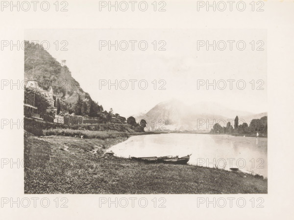 Street in Remagen, page, photomechanical print, title: 'Street in Remagen.', Remagen, c. 1889 - in or before 1899, Rhineland-Palatinate, paper, photoaquatint, height 132 mm x width 83 mm, street, A wide riverside scene in a faded sepia tone showing a grassy bank in the foreground with small rowboats pulled up at the water’s edge, a calm river stretching toward distant hazy hills and a pale sky, a wooded hillside on the left with ornate buildings partially visible among the trees and a small tower near the hilltop, a line of trees along the opposite shore and faint shapes of structures at the base of the distant hill.
