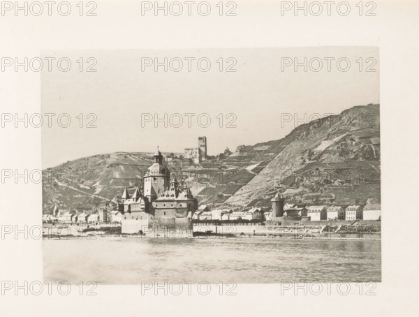 View of Kaub and Burg Pfalzgravenstein, page, photomechanical print, title: 'Caub and the Pfalz.', Rhine, c. 1889 - in or before 1899, paper, photoaquatint, height 83 mm x width 120 mm, city-view in general, 'veduta', timber framing, Rhineland-Palatinate, Kaub, Pfaffenporte **, A sepia-toned photograph showing a riverside town with a waterfront wall and a large stone building featuring turrets and a central dome rising above smaller pitched roofs; behind the town, terraced hills slope upward with a solitary square tower on a ridge and scattered vineyard-like patterns on the hillside; the river occupies the foreground with rippled water reflecting the shoreline, and the scene is framed by a wide pale border.