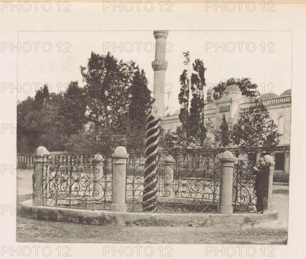 Serpentine Column in Constantinople, page, photomechanical print, title: 'Serpentine Column in the Hippodrome.', Constantinople, c. 1885 - in or before 1895, Ottoman Empire, paper, photoaquatint, height 94 mm x width 121 mm, column, pillar, architecture, Istanbul, Serpent Column, A black and white photograph shows an ornate wrought iron fence with swirling patterns supported by stone posts topped with rounded caps surrounding a tall twisted column. Behind the fence there are trees and a building with domed roof elements and arched windows, and a slender minaret rises above the foliage. A person stands near the fence on the right side, wearing a dark coat and hat and holding an object close to their face. The ground in front of the fenced area appears unpaved and textured.