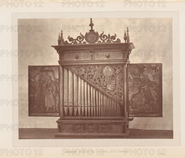 Organ of the South Kensington Museum in London, page, photomechanical print, inscription: 'Organ positive. German, XVI. Century.', London, c. 1869 - in or before 1874, paper, Woodburytype, height 116 mm x width 147 mm, organ, A photographic image of a wooden pipe organ with a decorative top featuring carved scrollwork and finials, a central circular medallion, a row of vertical organ pipes arranged in a triangular pattern, ornate carved panels with floral and figurative designs surrounding the pipes, and two hinged side panels displaying painted scenes of people and drapery, all set against a plain background and mounted on a carved base.