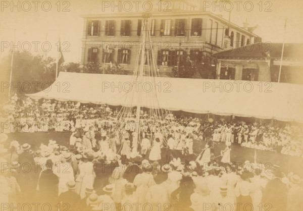 May-pole dance in Shanghai for the Diamond Jubilee of Victoria of the United Kingdom, The May-pole dance, photograph, page, photographer: WR Kahler (possibly), Shanghai, 1897, paper, albumen print, height 106 mm x width 151 mm, group dancing, commemoration, jubilee, festivities (wreath, garland, festive activities), A faded sepia photograph showing a large outdoor gathering in front of a multi-story building with shuttered windows and a covered awning; a tall pole with many ribbons extends upward from the center of the crowd where people in period clothing form a circle, while rows of spectators wearing hats fill the foreground and the area under the awning.