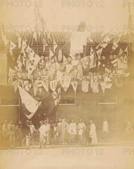 Mr. Satow's Photographic Establishment, Nanking Street, View of a photo studio presumably belonging to Erneste Mason Satow in Shanghai, the façade is decorated for the Diamond Jubilee of Victoria of the United Kingdom, photograph, page, photographer: WR Kahler (possibly), Shanghai, 1897, WR Kahler, photographer, paper, albumen print, height 150 mm x width 107 mm, commemoration, jubilee, festivities (wreath, garland, festive activities), A faded sepia photograph of a two-story building facade draped with multiple triangular and rectangular cloth flags and banners hanging from ropes across the upper level, with several large round decorative objects interspersed among the fabric, a large draped cloth hanging centrally from the balcony, and a row of tall windows visible beneath the decorations; a group of people stand in front of the building on the sidewalk, some wearing light-colored shirts and long garments, a few wearing hats, and one person holding a bicycle near the front, while the...