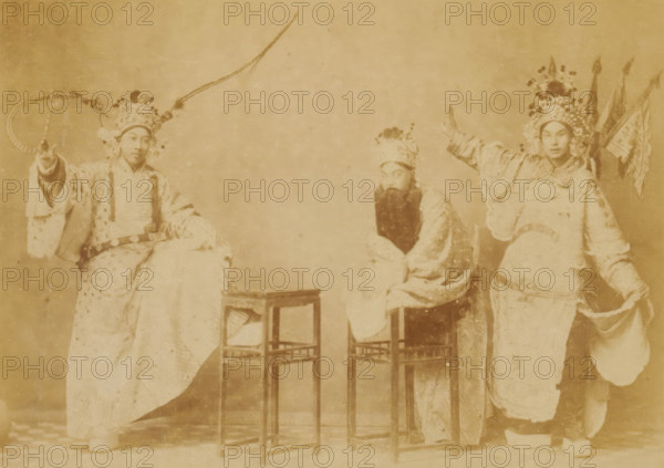 Portrait of three unknown Chinese men in theater costumes, Chinese theatrical, photograph, page, China, c. 1884 - in or before 1894, photographer, paper, albumen print, height 102 mm x width 144 mm, stage costume, A sepia-toned photograph mounted on paper shows three people in ornate traditional costumes and headdresses posed around two small wooden stools; the person on the left stands with one arm raised holding a circular prop and wears a long embroidered robe and a tall decorated headpiece with a long feather, the middle person sits slightly hunched with a long false beard and a patterned robe and headpiece, and the person on the right stands with one arm extended and wears a richly decorated robe, an elaborate floral headpiece, and holds several banners behind them; a decorative printed border surrounds the photograph on the mounting.