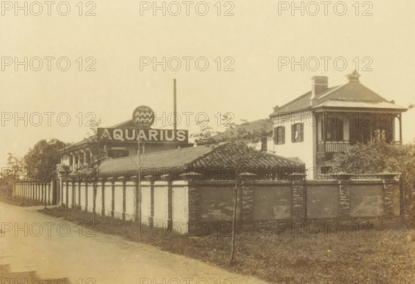 The Aquarius Company, Aquaris drinking water factory on Seward Road in Shanghai, photograph, page, Shanghai, c. 1884 - in or before 1894, photographer, paper, gelatin silver print, developed gelatin silver print, height 97 mm x width 140 mm, trade-company, A sepia photograph shows a street scene framed by a decorative border, featuring a long low wall with evenly spaced vertical sections and a tiled roof behind it, trees and shrubs scattered along the property, and a two-story building with a covered balcony, shuttered windows, and a hipped roof with a small roof ornament; a circular sign with a patterned emblem sits above the roofline near a taller vertical pole, and a paved road runs alongside the wall.