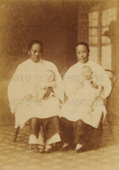 Portrait of two unknown Chinese nannies with babies on their laps, Chinese Amahs, photograph, page, China, c. 1884 - in or before 1894, photographer, paper, albumen print, height 144 mm x width 103 mm, baby-sitter, A faded sepia photograph bordered by decorative lines shows two seated women wearing light-colored tops and darker skirts, each holding an infant in white clothing on their laps; the women have similar hairstyles pulled back and neutral facial expressions, and the background includes a window with panes and a patterned floor beneath their feet.