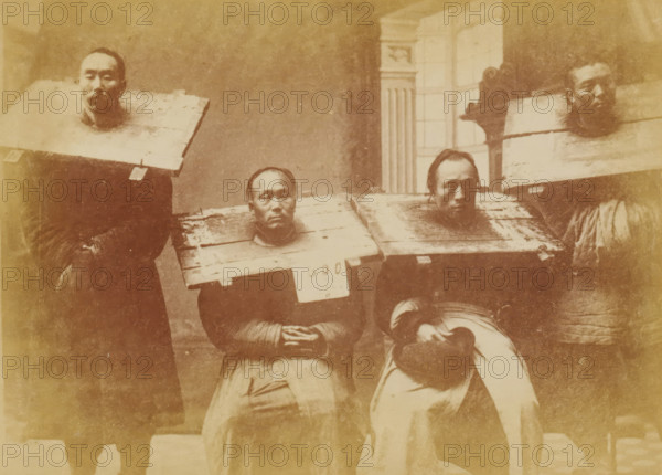 Portrait of four unknown Chinese men with a cangue around their necks, The cangue, photograph, page, China, c. 1884 - in or before 1894, photographer, paper, albumen print, height 104 mm x width 144 mm, Humiliation, A sepia-toned photographic print shows five seated and standing adults wearing wooden boards around their shoulders with rectangular openings framing their heads; the boards appear weathered with visible planks and fastenings, and two of the boards have small rectangular tags attached. The people display neutral facial expressions and are dressed in dark garments and layered clothing, with one person holding a brimmed hat in their lap. The background includes a wall and a tall window with mullions and a decorative vertical architectural element, and the image is set within an ornate printed border on aged paper.