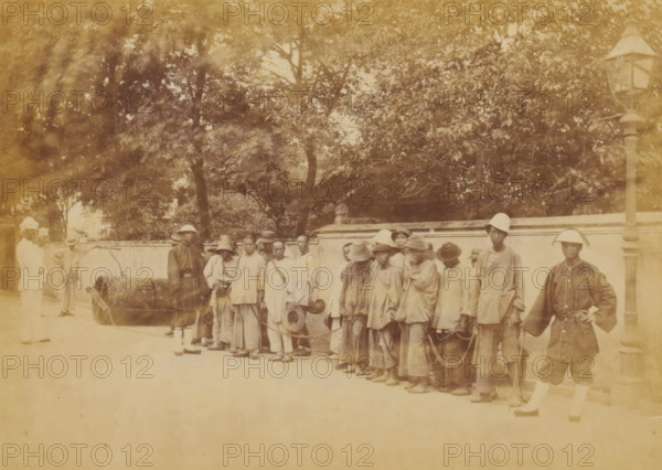 Group of Chinese people chained together under the supervision of guards, The chain-gang, photograph, page, China, c. 1884 - in or before 1894, photographer, paper, albumen print, height 102 mm x width 144 mm, workhouse, penal camp, penitentiary, chain gang, A sepia-toned photograph mounted on a decorative bordered page shows a group of people standing in a line along a street in front of a low wall with trees behind it; most individuals wear loose clothing and hats, and several are connected by chains; two men in lighter uniforms stand near a large cylindrical object on the left, while a man in a darker uniform stands by a tall streetlamp on the right; the scene includes a paved ground and dappled light from the foliage above.