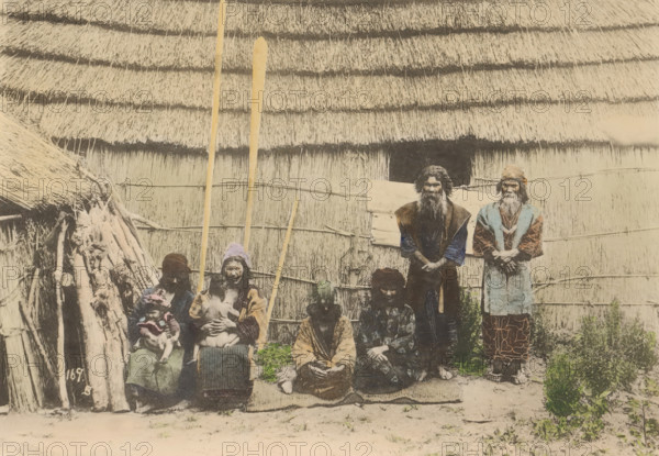 Ainues, the Native of Hokkaido, Japan, Portrait of unknown people of the Aino in front of a house in Japan, page, photomechanical print, number: '169.B', maker: Kozaburo Tamamura (possibly), c. 1895 - c. 1905, paper, collotype, photoprint, hand colored, height 105 mm x width 151 mm, human types, peoples and nationalities, A faded photograph shows a group of people posed in front of a large thatched wall and roof made of layered straw. Several tall wooden poles lean upright against the wall. Two older men stand at the back with long beards and layered clothing, one wearing a patterned garment and the other wearing a vest over darker sleeves. In front of them three seated adults wear varied patterned and textured clothing, one holding a small child and another holding an infant; one seated person has a head covering. To the left a simple structure of sticks and thatch is visible with bundled wood and rough supports. The ground in front of the group is bare earth with patches of low veget...