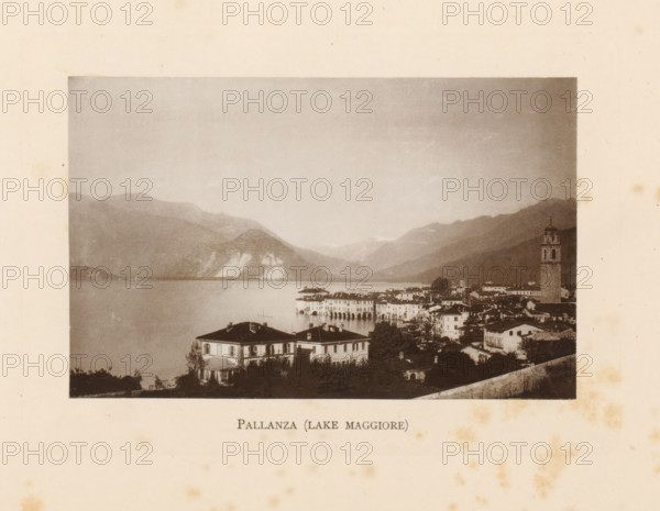 View of Pallanza on Lake Maggiore in Italy, Pallanza (Lake Maggiore), page, photomechanical print, Italy, c. 1874 - in or before 1884, paper, collotype, photoprint, height 86 mm x width 136 mm, lake, prospect of village, silhouette of village, mountains, Piemonte, Verbania, A sepia-toned photograph framed on aged paper shows a lakeside town with clustered buildings and a prominent bell tower rising above the rooftops, a waterfront with arched colonnades extending into the water, and villas with shuttered windows and striped awnings in the foreground; calm water fills the middle ground and reflects the shoreline, while layered hills and mountains fade into the sky in the background.