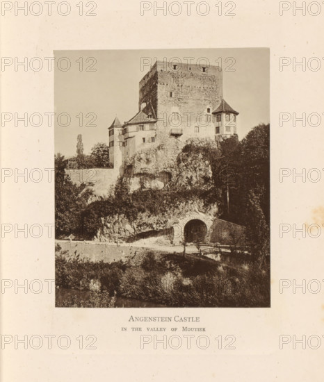 View of Schloss Angenstein, Angenstein Castle in the Valley of Moutier, page, photomechanical print, Basel-Landschaft, c. 1874 - in or before 1884, paper, collotype, photoprint, height 120 mm x width 100 mm, castle, Schloss Angenstein, A monochrome photograph of a large stone castle perched on a rocky hill, showing a tall rectangular keep with small windows and rough masonry, flanked by smaller turrets with conical roofs and multiple windowed facades; vegetation and trees grow around the rocky base, and a rounded arched opening is visible at the rock face leading into a tunnel or passage; a low stone wall and a path run along the hillside below the castle, and the scene is framed by a wide blank border.
