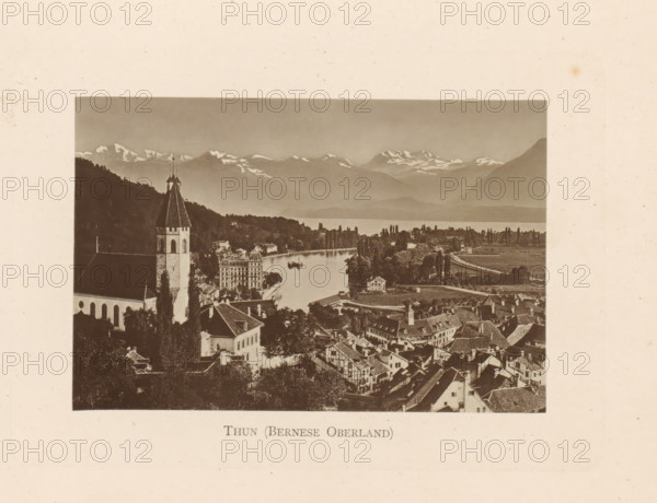 Thun (Bernese Oberland), View of Thun in Switzerland, page, photomechanical print, Switzerland, c. 1874 - in or before 1884, paper, collotype, photoprint, height 85 mm x width 128 mm, city-view in general, 'veduta', mountain-range, snow peaks, A sepia photograph showing a lakeside town with clustered rooftops and narrow streets in the foreground, a tall church tower with a steep pointed roof rising among trees to the left, a river or inlet curving through the middle of the scene with boats on the water, rows of trees along the shoreline and open fields beyond, and distant snow-capped mountains under a clear sky.