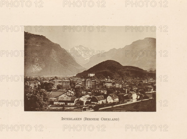 Interlaken (Bernese Oberland), View of Interlaken in Switzerland, page, photomechanical print, Switzerland, c. 1874 - in or before 1884, paper, collotype, photoprint, height 85 mm x width 130 mm, city-view in general, 'veduta', mountain-range, snow peaks, A sepia-toned rectangular photograph shows a town at the base of forested hills and distant snow-capped mountains, with clustered buildings including multi-story structures and smaller houses with pitched roofs, trees interspersed among them, and a road visible along the foreground.