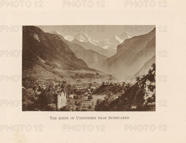 The ruins of Unspunnen near Interlaken, View of the ruins of Burg Unspunnen near Interlaken, page, photomechanical print, Switzerland, c. 1874 - in or before 1884, paper, collotype, photoprint, height 82 mm x width 127 mm, mountains, ruin of a dwelling, house, castle, A sepia-toned landscape photograph framed by a wide margin shows a valley between steep, dark mountain slopes with snow-capped peaks visible in the distance. In the foreground there are scattered trees, open fields, and several small buildings, including a prominent ruined stone structure on a rise and a house perched on a hill. Layers of ridges recede toward the distant glaciers and peaks, and the scene has a soft, hazy atmospheric gradation from the foreground to the distant mountains.
