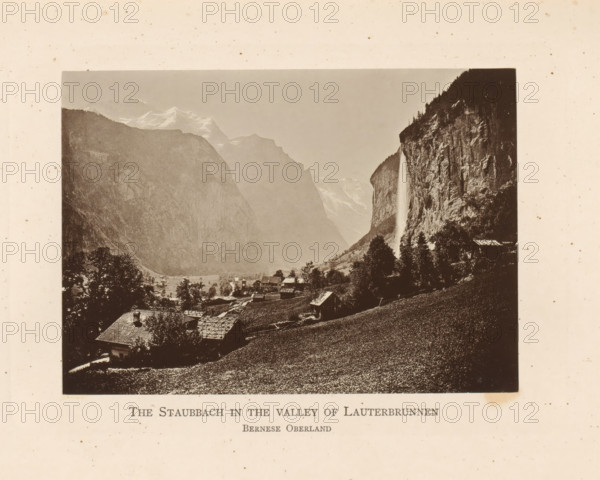 View of the Lauterbrunnen Valley with the Staubbach Waterfall, The Staubbach in the Valley of Lauterbrunnen Bernese Oberland, page, photomechanical print, Bern (canton), c. 1874 - in or before 1884, paper, collotype, photoprint, height 93 mm x width 131 mm, waterfall, dale, valley, Lauterbrunnen, Staubbachwaterval, A sepia-toned photograph shows a valley framed by steep rocky cliffs and distant snow-capped peaks under a pale sky, with a tall narrow waterfall cascading down a cliff face on one side; below, clustered rustic buildings with pitched roofs sit among trees and sloping grassy fields, and a foreground hillside covered in textured vegetation slopes toward the village.