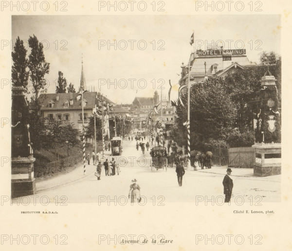 View of the Avenue de la Gare in Fribourg, Avenue de la Gare, page, photomechanical print, photographer: Ernest Lorson, Fribourg, maker: SADAG, 1897, paper, collotype, photoprint, height 113 mm x width 163 mm, tram, street, advertising, A wide street scene showing a central road lined with tall trees and multi-story buildings, a steep-roofed building with a spire visible in the background, and a large hotel building with a rooftop sign and a flagpole on the right; the street is busy with pedestrians dressed in period clothing, horse-drawn carriages and trams, lampposts and decorative poles with flags along the sidewalks, a woman walking toward the foreground near the center, and a man standing at the right edge, with stone pillars and fencing visible near the sidewalks and dense foliage on the right side.