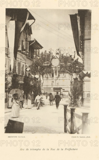 Arc de triomphe de la Rue de la Préfecture, View of a street with an arch of honor in Fribourg, page, photomechanical print, maker: Ernest Lorson, Fribourg, maker: SADAG, 1897, paper, collotype, photoprint, height 162 mm x width 111 mm, street, triumphal arch, Friborg, A sepia-toned street scene framed by buildings on both sides shows an archway decorated with foliage and draped fabric spanning the road, with hanging round ornaments and flags. People in period clothing walk and stand beneath and near the arch, including women in long dresses and a man wearing a uniform with a hat. Shuttered windows and awnings are visible on the façades, and lamp fixtures and potted greenery adorn the street. A wooden railing runs along the right foreground, and a multi-story building with shuttered windows fills the background behind the arch.