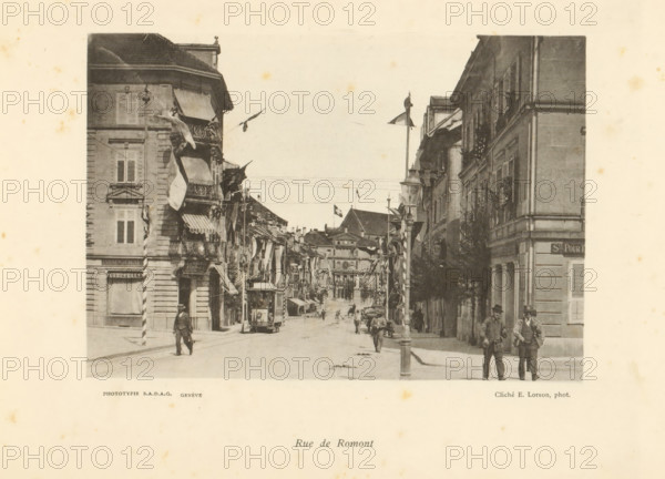 Rue de Romont, View of Rue de Romont in Fribourg, page, photomechanical print, photographer: Ernest Lorson, Fribourg, maker: SADAG, Genève, 1897, paper, collotype, photoprint, height 113 mm x width 162 mm, tram, street, advertising, A monochrome street scene framed by tall buildings on both sides, with awnings and balconies projecting over the sidewalk and flags hanging from several facades, a tram car on tracks near the foreground, pedestrians walking along the street and standing on the sidewalks, a lamppost or pole rising near the center of the image, architectural details such as shuttered windows and decorative stonework on the buildings, distant buildings visible down the street and a sky above.