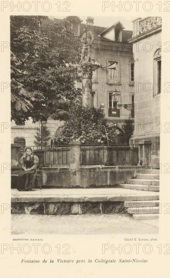 View of a fountain in Fribourg, Fontaine de la Victoire près la Collégiale Saint-Nicolas, page, photomechanical print, maker: Ernest Lorson, Fribourg, maker: SADAG, 1897, paper, collotype, photoprint, height 162 mm x width 110 mm, ornamental fountain, Friborg, A black and white photograph showing a stone fountain area surrounded by a low stone wall and metal railing, with shrubbery and a tall sculpted column topped by a standing statue rising behind the enclosure; to the left a large tree with dense foliage partially obscures the background buildings, which include multi-storey façades with windows and a decorative cornice visible on the right; a woman in period dress sits on a bench in front of the stone wall at the left side of the image, and stone steps lead up to the fountain area from the right foreground.