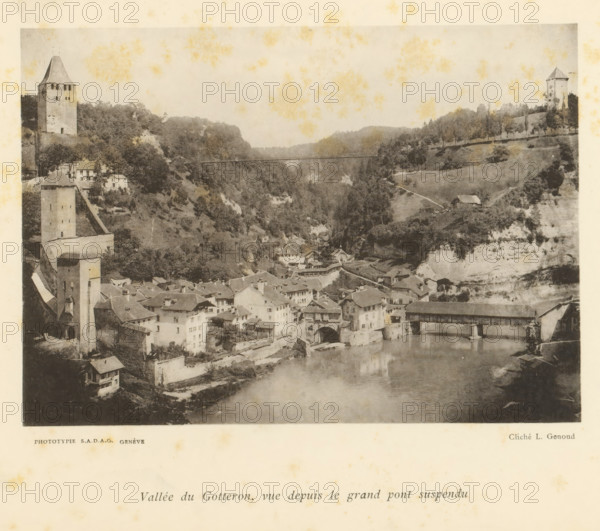 Vallée du Gotteron, vue depuis le grand pont suspendu, View of the Vallée du Gottéron from the grand pont suspendu in Fribourg, page, photomechanical print, maker: L. Genoud, Fribourg, maker: SADAG, Geneva, 1897, paper, collotype, photoprint, height 113 mm x width 157 mm, city-view, and landscape with man-made constructions, dale, valley, A sepia-toned photograph showing a valley with a river in the foreground and a clustered village of stone buildings with pitched roofs along its banks, a covered wooden bridge crossing the river, several tall stone towers on the hillsides, terraced slopes and trees behind the village, a narrow suspension bridge spanning the gorge in the distance, and a hillside road leading to a small isolated building.