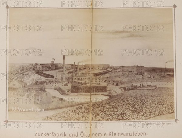 View of a sugar factory in Klein Wanzleben, Zuckerfabrik und Ökonomie Kleinwanzleben, photograph, page, photographer: Wilhelm Höffert, Klein Wanzleben, c. 1884 - in or before 1894, Saxony-Anhalt, paper, albumen print, height 129 mm x width 189 mm, Photo on frontispiece., factory in landscape, sugar, façade (of house or building), A wide sepia-toned panoramic image of an industrial complex set in a rural landscape, featuring multiple long low buildings and larger multi-story factory structures, tall smokestacks emitting plumes of smoke, rows of smaller dwellings along a foreground road, fenced yards and open storage areas, a body of water with adjacent scaffolding or framed construction, terraces of cultivated fields and dense vegetation on rolling ground, scattered trees and distant low hills under a pale sky with faint cloud formations, and a decorative border framing the scene.