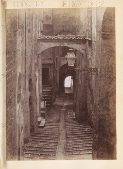View of a street, presumably in Italy, photograph, page, Italy, c. 1850 - in or before 1860, photographer, paper, albumen print, height 134 mm x width 97 mm, street, A narrow, tiled street flanked by tall, weathered stone walls and buildings, with an arched passageway spanning the alley near the top. A decorative metal bracket holds a lantern projecting into the space below the arch. The ground is covered with rows of curved roof tiles forming a pathway that leads toward a darker archway in the distance. Several small windows and recessed doorways are set into the walls, and a suspended light fixture is visible further down the alley. The surfaces show textured stone, patches of wear, and subtle shadowing along the vertical facades.