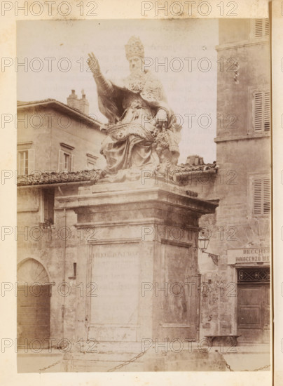 Statue of Pope Julius III in Perugia, photograph, page, Perugia (city), c. 1850 - in or before 1860, photographer, Umbria, paper, albumen print, height 134 mm x width 97 mm, Person, Julius III, monument, statue, A sepia-toned photograph showing a large stone statue of a seated robed figure wearing an ornate headdress and raising one hand, positioned atop a tall rectangular pedestal. The statue displays detailed drapery and decorative elements, and a sculpted head or bust rests near the figure's side. Surrounding the monument are aged stone buildings with shuttered windows, tiled roofs, a small balcony with a potted plant, and an arched doorway. A wall-mounted lantern projects from one building, and a section of chained bollard is visible at the base of the pedestal. The image has a faded, slightly stained border and overall worn texture.
