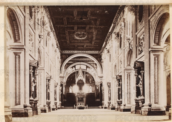 Interior of St. John Lateran in Rome, photograph, page, St. John Lateran, c. 1850 - in or before 1860, photographer, Rome, paper, albumen print, height 96 mm x width 136 mm, interior of church, A wide interior view of a grand hall with a high coffered ceiling featuring a circular decorative medallion, flanked by tall ornate walls with arched niches and pilasters; the walls display sculpted reliefs, carved figures in recessed alcoves, and decorative panels, with ornate capitals and cornices. A large arched opening leads to a raised focal area containing an intricately carved canopy or altar-like structure with vertical decorative elements and an elaborate frontal panel, and the floor in front is open and unobstructed. The image is monochrome, showing strong contrasts between the dark ceiling and lighter stonework, and the scene is symmetrical with repeated architectural details along both sides.