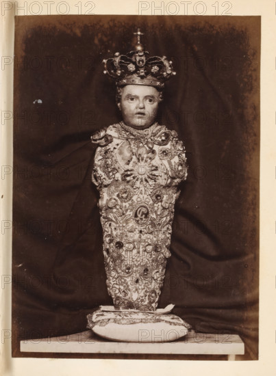 Wax figure of a baby with a crown and decorated with gemstones, photograph, page, c. 1850 - in or before 1860, photographer, paper, albumen print, height 135 mm x width 97 mm, piece of sculpture, reproduction of a piece of sculpture, A vertically oriented sepia photograph showing a single bustlike figure centered against a dark draped background, wearing an ornate crown with rounded arches and decorative elements, a lifelike face with neutral expression, and a heavily embellished garment covered in intricate embroidery, jewels, and medallionlike ornaments; the figure rests on a decorative cushion placed on a flat surface, and the image is bordered by a lighter margin.