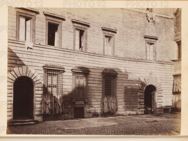 View of the facade of probably Palazzo Cenci in Rome, photograph, page, Rome, c. 1850 - in or before 1860, photographer, paper, albumen print, height 98 mm x width 133 mm, hôtel, ie small palace in city, A sepia-toned photograph of a stone building facade with a cobblestone street in front, showing an upper row of rectangular windows with decorative frames and an open window with a small figure leaning on the sill, and a lower row featuring arched doorways and rectangular windows protected by metal grilles with draped fabric hanging beneath them; the wall surface displays horizontal stone courses and weathered texture, and there are shadows cast across the facade and on the ground.