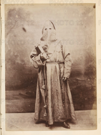 Portrait of someone in a monk's habit and with a hood over their head, photograph, page, Italy, c. 1850 - in or before 1860, photographer, paper, albumen print, height 136 mm x width 98 mm, monks and nuns, A sepia-toned photograph of a person standing against a plain backdrop wearing a full-length hooded robe with a pointed hood that covers the face except for two eye openings, a belt tied at the waist with a cord and hanging tassels, a strand of beads hanging from the waist, and a circular patch on the chest; the person holds a rectangular object in one hand and a staff or pole in the other, and wears dark shoes visible beneath the hem of the robe.