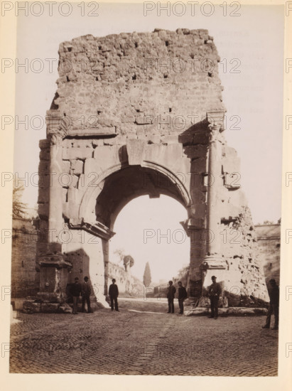 View of an ancient city gate of Rome, photograph, page, Rome, c. 1850 - in or before 1860, photographer, paper, albumen print, height 134 mm x width 97 mm, city-gate, A large ancient stone archway with weathered blocks and columns fills the frame, showing a rounded arch opening and a rough textured upper wall. The arch rests on thick supports with visible carvings and broken edges, and fragments of stone and rubble sit at the base. A cobbled street stretches beneath the arch, leading to a distant tree visible through the opening. Several men in period clothing and hats stand and pose near the arch and along the sides, some leaning against the stone and others standing on the pavement. The lighting casts shadows across the arch and the street, and portions of adjoining masonry walls are visible on either side of the structure.