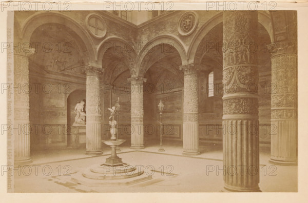 Firenze. Cortile di Palazzo Vecchio, riordinato nel 1434 da Michelozzi, Courtyard of the Palazzo Vecchio in Florence, photograph, page, number: '3036.', photographer: Edizioni Brogi (possibly), Palazzo Vecchio, c. 1878 - in or before 1888, Florence, Tuscany, paper, albumen print, height 94 mm x width 147 mm, inner court of palace or 'hôtel', A vaulted interior with multiple decorated columns supporting rounded arches, a central raised fountain with a pedestal and basin, ornate carved column capitals and patterned column shafts, a sculpted figure in an alcove to the left, two tall lamppost-like fixtures near the fountain, wall panels and painted or carved arch surfaces, a small window letting light through on the right, and a patterned floor with stepped circular base beneath the fountain.