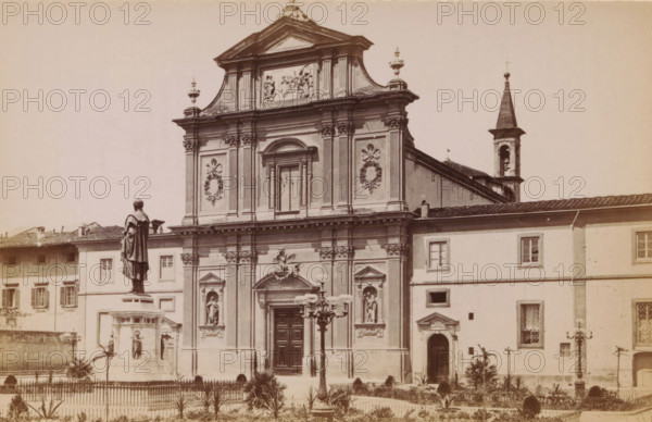 Firenze. Facciata della chiesa di S. Marco, opera dei 1780 di Giovacchino Pronti, Exterior of the Basilica di San Marco in Florence, photograph, page, photographer: Edizioni Brogi (possibly), Florence, c. 1878 - in or before 1888, Tuscany, paper, albumen print, height 92 mm x width 148 mm, church (exterior), facade (of house or building), San Marco, A sepia-toned photograph of a large ornate church façade with decorative columns, sculpted reliefs, and statues set in niches, topped by a triangular pediment and ornamental urns, flanked on the right by a bell tower with a pointed spire; in front of the church a tall statue of a robed figure stands on a multi-tiered pedestal within a fenced garden area containing small plants and lampposts, and adjacent buildings with shuttered windows form the background.
