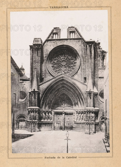 Fachada de la Catedral, Facade of the Cathedral of Tarragona, Tarragona, page, photomechanical print, c. 1874 - in or before 1884, Catalonia, paper, collotype, photoprint, height 160 mm x width 111 mm, church (exterior), grave, tomb, Tarragona Cathedral, A black and white photograph of a cathedral façade showing a large circular rose window above a pointed arch entrance, flanked by tall stone pillars and ornate sculpted figures on both sides of the doorway, with decorative carvings and niches visible on the lower façade, a streetlamp standing in front of the entrance, adjacent buildings partially visible at the sides, and the image bordered by a textured paper frame.
