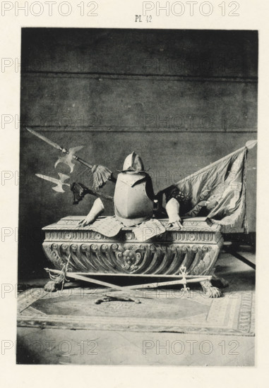 Sarcophagus with armor and weapons, page, photomechanical print, number: 'Pl 42', Barcelona (city), c. 1880 - in or before 1881, Catalonia, paper, collotype, photoprint, height 147 mm x width 103 mm, weapons, sarcophagus, harness, A photographic print shows a decorative stone or carved base with scroll and floral motifs resting on lion paw feet, atop which sits a suit of armor including a breastplate and a helmet with a brim. Pole weapons with ornate heads and tassels are arranged behind and across the armor, and a draped fabric flag with visible folds is positioned to one side. In front of the base, crossed swords rest on the floor over a patterned rug with a central medallion design.