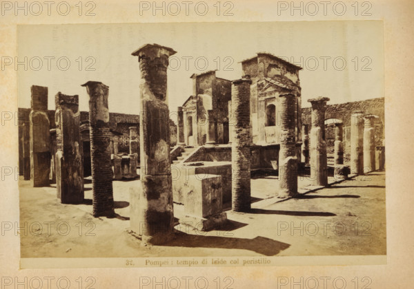 Pompeii: tempio di Iside col peristilio, Ruin of the Tempio di Iside in Pompeii, photograph, page, number: '32.', c. 1878 - in or before 1888, photographer, paper, albumen print, height 103 mm x width 153 mm, ruin of a building, architecture, temple, shrine, Roman religion, Tempio di Iside, A sepia photograph shows a sunlit archaeological site with rows of weathered stone columns and rectangular stone blocks arranged on a sandy ground, some columns topped with rough capitals and others with flat fragments, casting long shadows; in the background there are ruined stone walls and a partially intact façade featuring an arched opening and decorative pediment, with worn brick and plaster textures visible across the structures.