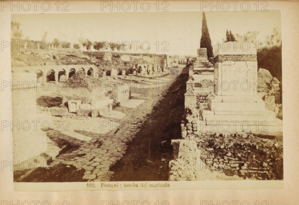 Pompeii: tomba del marinaio, Ruin of tombs at Pompeii, photograph, page, number: '102.', c. 1878 - in or before 1888, photographer, paper, albumen print, height 103 mm x width 156 mm, ruin of a building, architecture, grave, tomb, A sepia-toned photograph showing a ruined stone roadway flanked by ancient masonry and larger stone monuments on the right, with a sequence of arched openings and low stone structures on the left; the road surface displays irregular paving stones and a darker strip running along its center, leading toward distant ruins and a row of trees on a raised embankment; rubble, broken blocks, and fragmented walls populate the foreground and middle distance, and a tall cypress-like tree rises near the right-hand monuments against a pale sky.