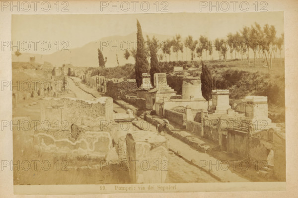 Pompeii: via dei Sepolcri, Via dei Sepolcri in Pompeii, photograph, page, number: '99.', c. 1878 - in or before 1888, photographer, paper, albumen print, height 101 mm x width 153 mm, public road, ruin of a building, architecture, grave, tomb, A sepia-toned view of an ancient street flanked by low stone walls and ruined structures, with rectangular stone tombs and raised platforms lining one side and carved facades and masonry remains on the other, a narrow paved roadway running between them, tall cypress trees rising above the ruins and a row of slender trees on a ridge in the background, distant hazy hills beyond, and uneven ground and scattered stones in the foreground.