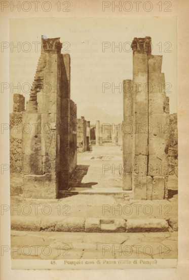 Ruin of the Casa di Pansa in Pompeii, Pompei: casa di Pansa (Edile di Pompei), photograph, page, number: '48.', Pompeii, c. 1878 - in or before 1888, photographer, paper, albumen print, height 153 mm x width 103 mm, ruin of a building, architecture, housing, A sepia-toned photograph showing a narrow ruined corridor framed by tall stone pillars and walls with visible blocks and weathered surfaces, decorative capitals atop two pillars at the near ends, a stepped broken wall on the left, a flat paved foreground with worn stones, shadows cast along the passage, and additional distant columns and wall fragments visible down the central walkway.