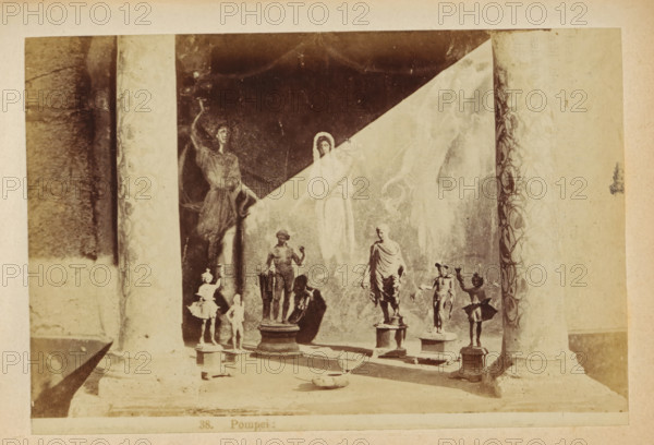 Pompeii: Sculptures at Pompeii, photograph, page, number: '38.', c. 1878 - in or before 1888, photographer, paper, albumen print, height 103 mm x width 153 mm, sculpture, ruin of a building, architecture, A faded photograph shows a shallow display area framed by two textured vertical columns with decorative borders, inside which several small statues stand on pedestals and plinths arranged along a low platform; the statues vary in pose and dress, including a draped figure, a standing nude figure, and smaller figures with outstretched arms, while a painted backdrop behind them features a diagonal light area and faint ghostly figures including two larger painted human forms, one with an arm raised and another with a draped garment and halo-like outline, and the overall scene has a worn, sepia-toned appearance with shadows falling across the foreground.