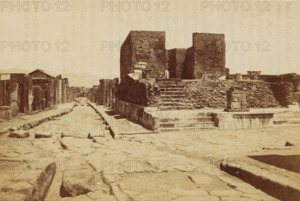 View of the ruins of the Tempio della Fortuna Augusta in Pompeii, Pompei, fortune magasin terres cuites temple fortune Statue a Ciceron fouilles, photograph, page, number: '3010', Pompeii, c. 1878 - in or before 1888, photographer, paper, albumen print, height 104 mm x width 143 mm, landscape with ruins, temple, shrine, Roman religion, Tempio della Fortuna Augusta, A wide view of an ancient street paved with large irregular stones leading into the distance between rows of low stone ruins and facades, with a raised stone platform and stepped structure on the right made of rough blocks and a taller rectangular stone building behind it, shallow gutters or channels running along the street edges, scattered stone blocks and fragments on the pavement, worn steps and a small cylindrical stone feature near the platform, and a pale sky above.