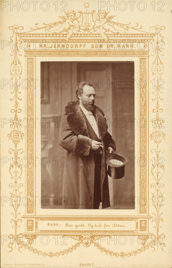 Portrait of the actor Peter Jerndorff as Doctor Rank in A Doll's House, Hr. Jerndorff sum Dr. Rank, photograph, page, inscription: 'Rank:, Ilden.', photographer: JA Schulz, printer (person): Edvin Jantzen, publisher: Ernst Bojesen, c. 1870 - in or before 1880, JA Schulz, Edvin Jantzen, Ernst Bojesen, photographer, printer (person), publisher, paper, albumen print, albumendruk, height 99 mm x width 62 mm, Person, Peter Jerndorff, portrait of actor, actress, A sepia-toned framed photograph shows a man standing indoors wearing a long coat with a fur collar and fur cuffs, a suit underneath, and a bow tie; he has a beard and mustache and is holding a top hat in one hand while the other hand holds a small object near his waist; the decorative frame around the photograph features symmetrical ornamental flourishes, cherub figures at the top, and intricate border patterns on a light background.